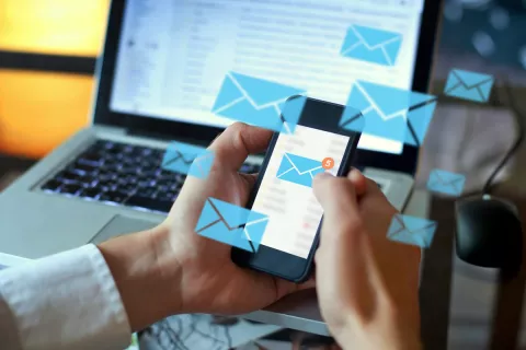 Zwei Hände halten ein Smartphone, auf dem mehrere Newsletter-Nachrichten ankommen. Im Hintergrund ist ein offener Laptop mit E-Mails.